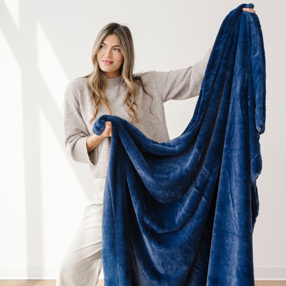 A woman holds up a deep blue lush Saranoni blanket that is a luxury blanket. This cozy blanket is a soft throw blanket and a faux fur blanket. This soft blanket is great as a home decor staple, comfort gift, or everyday relaxation item. Enjoy this Saranoni blanket for years to come.
