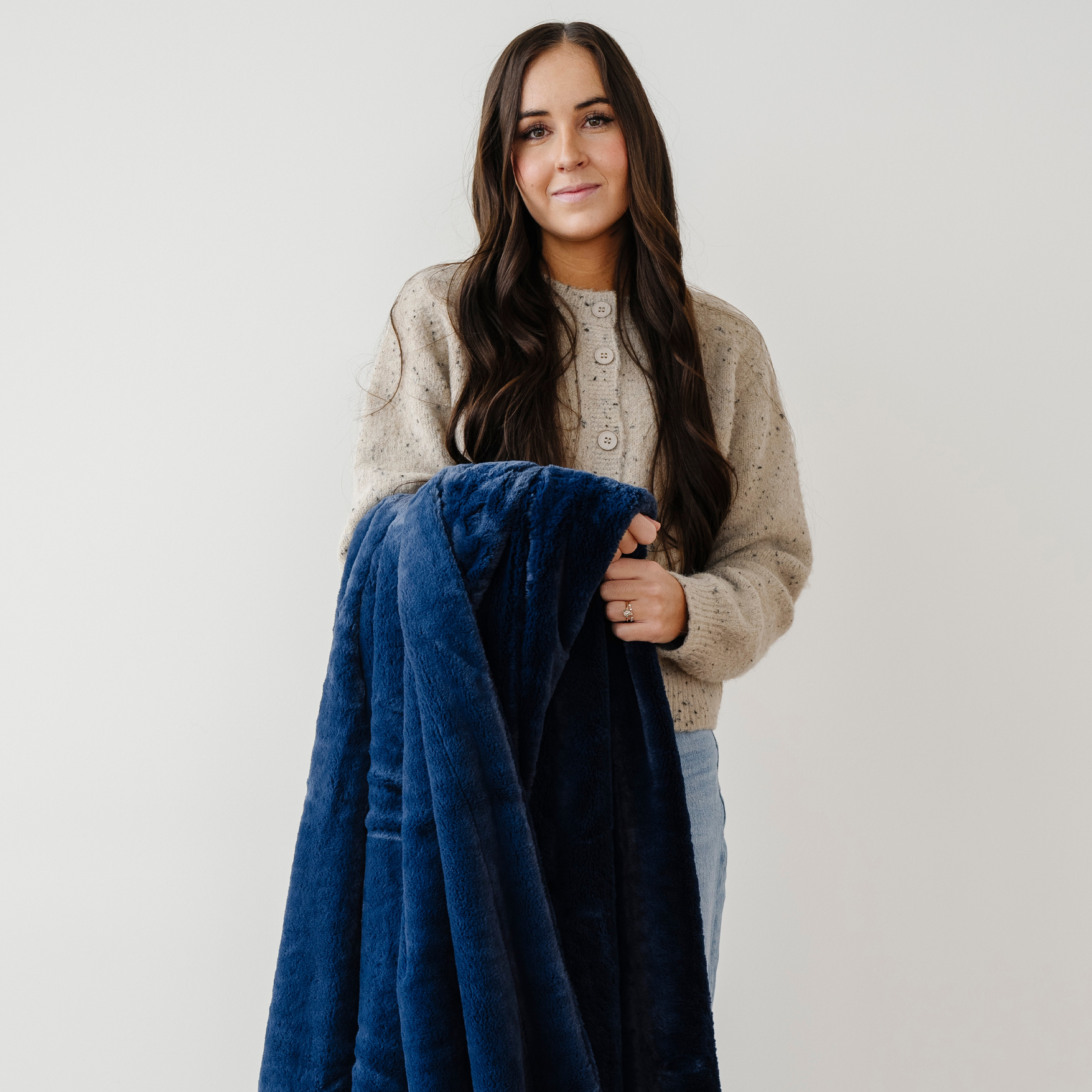 A woman is holding a deep blue lush Saranoni blanket. This luxury blanket is a soft throw blanket and a faux fur blanket. This soft blanket is great as a home decor staple, comfort gift, or everyday relaxation item. Elevate your home decor with this faux fur blanket. Enjoy this cozy blanket for years to come.