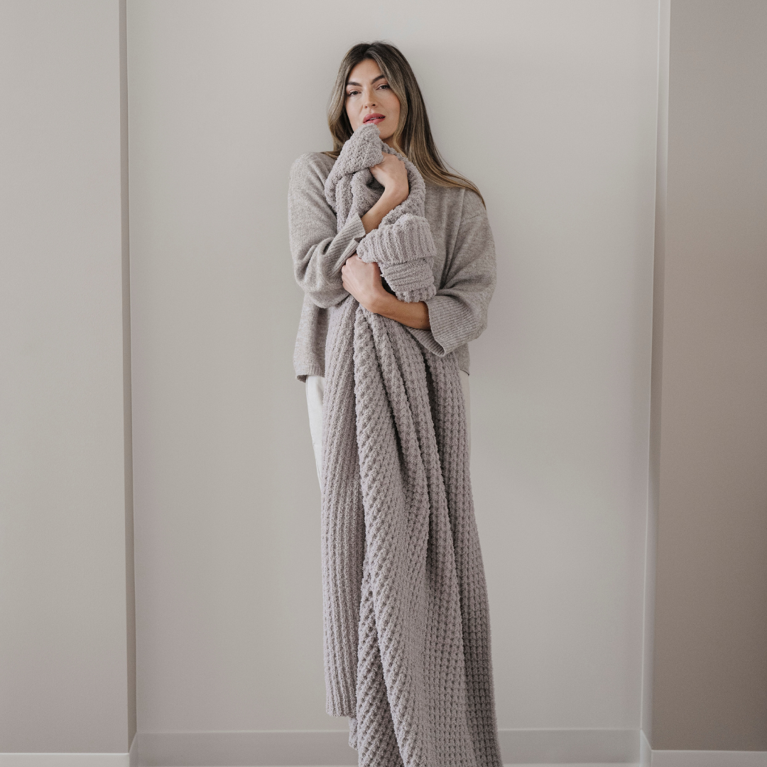A woman holds a light tan blanket that is a Saranoni blanket and is very stretchy and breathable. Use this cozy blanket for gifting, relaxing, or as home decor. Enjoy this luxurious blanket for years to come