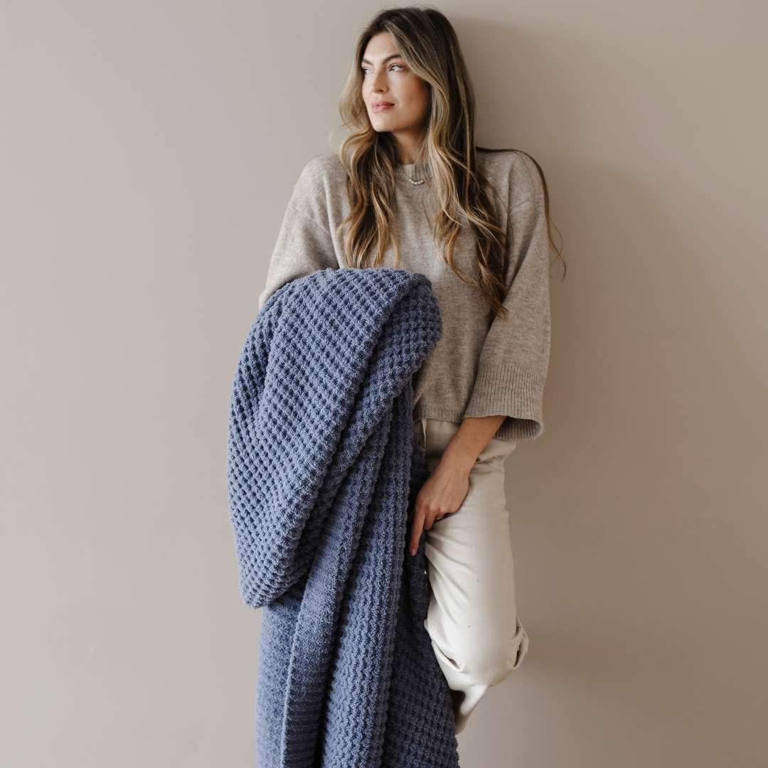 A woman is holding a Saranoni blanket that is a blue waffle knit blanket. This soft blanket is a luxury blanket and a Saranoni blanket. Use this blanket as a comfort gift, relaxation blanket, or home decor. 