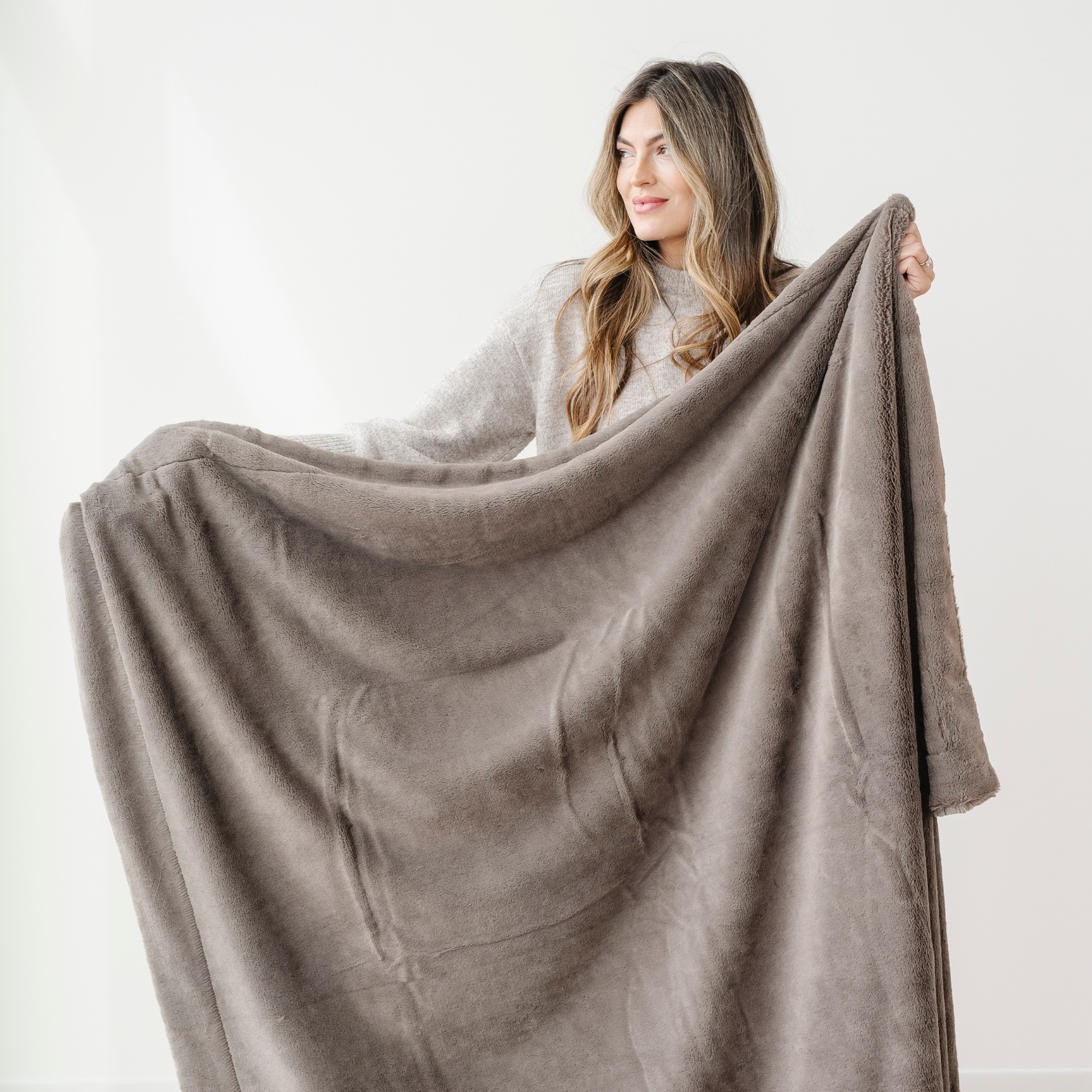A woman smiles and holds up a brown blanket that is a lush Saranoni blanket. This luxury blanket is a soft blanket and is super cozy. This faux fur blanket is great for gifting, relaxation, or as an addition to your home decor..