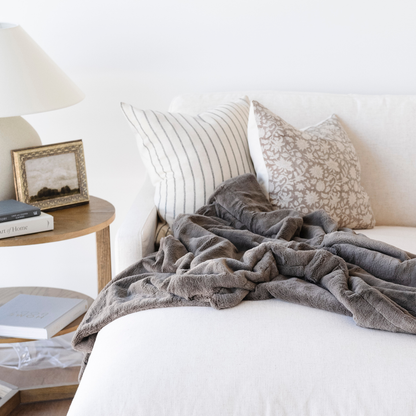 A brown lush Saranoni blanket is draped on a couch. This soft blanket is a luxury throw blanket and a faux fur blanket. This soft blanket is great as a home decor staple, comfort gift, or everyday relaxation item. Enjoy this Saranoni blanket for years to come.