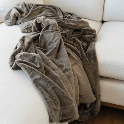A brown blanket that is a lush blanket is beautifully draped on a couch. This soft blanket is a Saranoni blanket and a luxury blanket. This cozy blanket is great as a home decor staple, everyday comfort item, or as a comfort gift. Elevate your home decor with this faux fur blanket that is an extra large blanket. 
