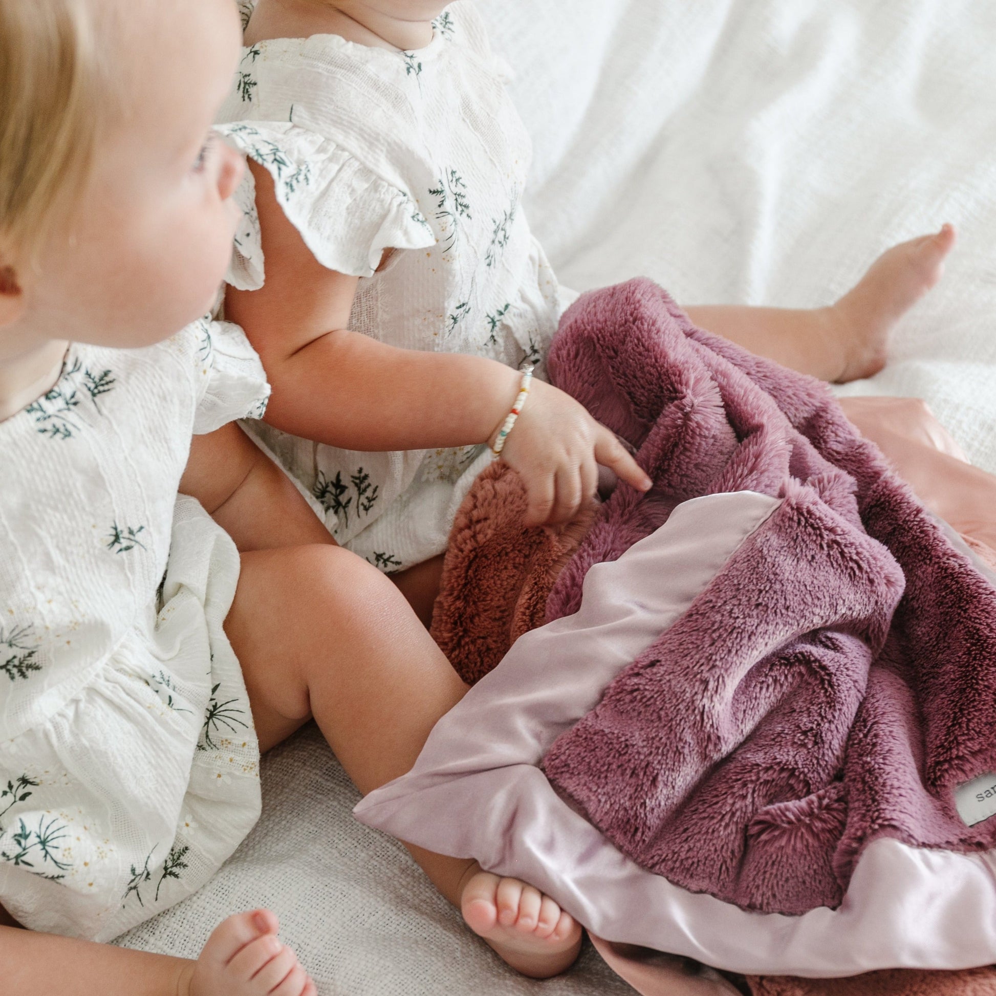 Two babies lay with a beautiful dusty purple colored satin blanket.