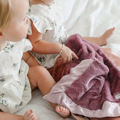Two babies lay with a beautiful dusty purple colored satin blanket.