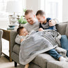 Two brothers and a dad laugh and lay together under a beautiful grey faux fur blanket.