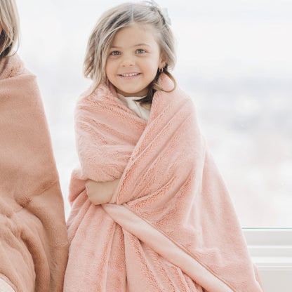 A little girl has wrapped around her a Blossom (light peachy pink) Colored Lush Saranoni Blanket. The soft blanket is a small blanket and a baby blanket or toddler blanket.