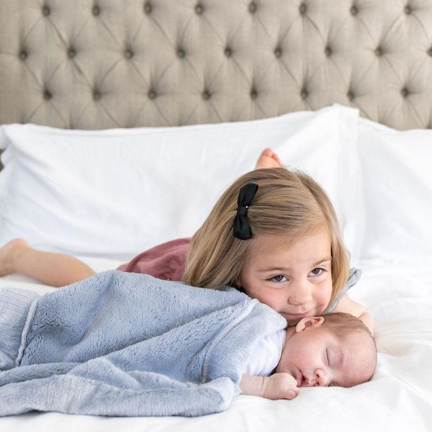 A little girl lays by her baby brother who is wrapped in a light blue soft mini Saranoni Baby Blanket. The Saranoni Blanket is the Lush Fabric and the specific color is called Storm Cloud. The blanket is a small blanket and a mini blanket.