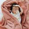 Clay@A baby with a pacifier in her mouth lays comfortably in a warm coral colored blanket.