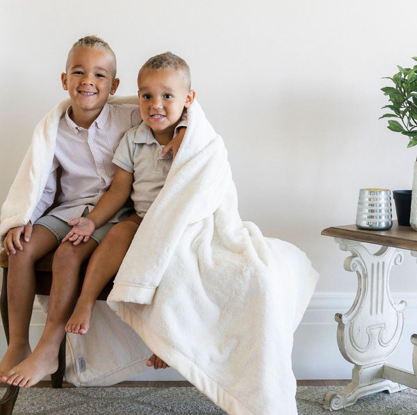Two brothers sitting on a bench wrapped in a white full sized blanket