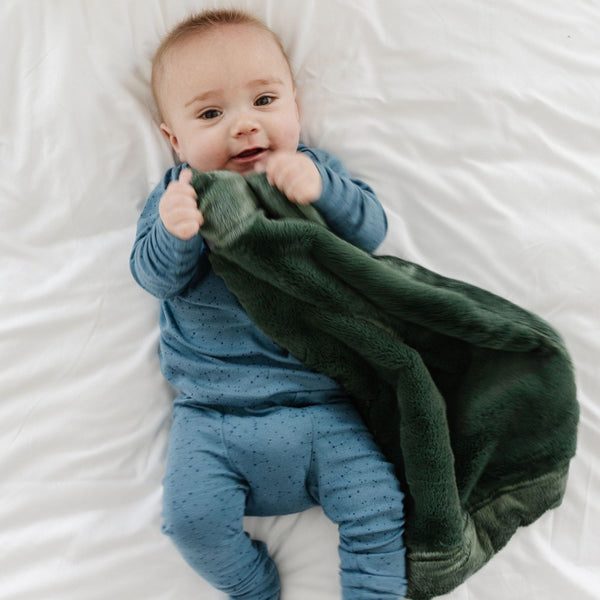 A baby lays on a bed while holding his dark green infant lush blanket.