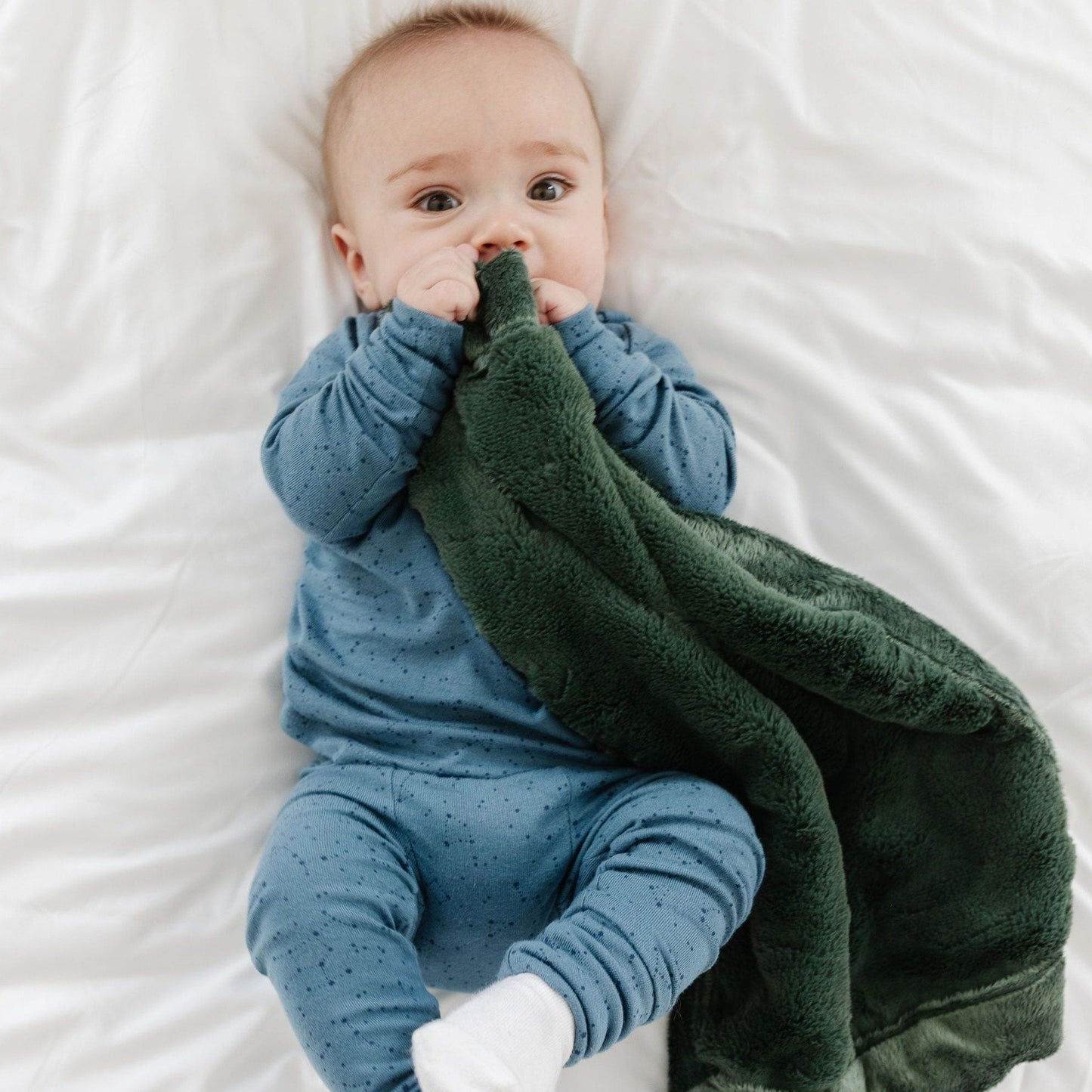 A baby boy laying on a bed holds a mini Hunter Green Colored Lush Saranoni Blanket. The soft blanket is a small blanket and a baby blanket or toddler blanket.