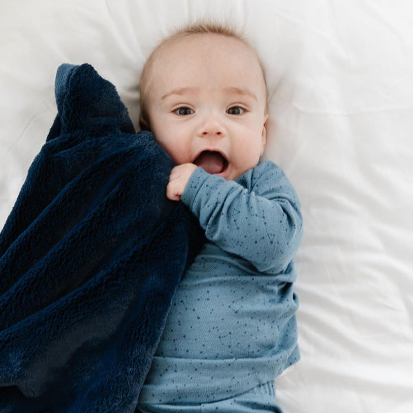 A cute baby with his mouth wide open holds a cozy dark blue blanket up to his face.