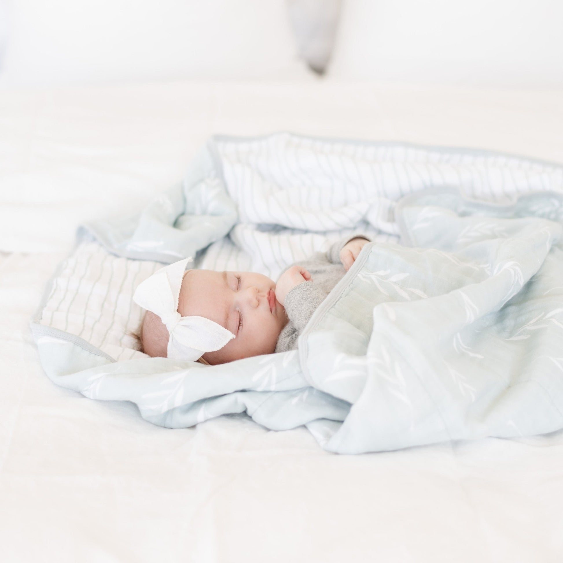 A baby girl sleeps wrapped in a cotton muslin quilt. This soft blanket is light blue and white with a floral and striped pattern. This blanket is also a Saranoni blanket and it's cute pattern makes it a great baby gift or toddler gift. 