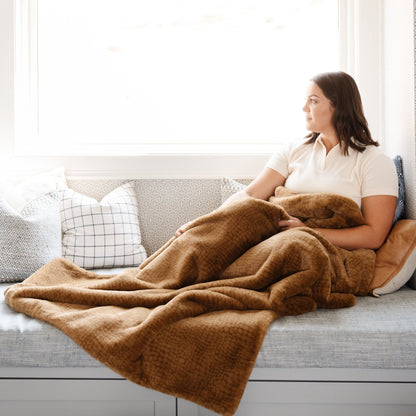 A woman sits with a Saranoni Grand Faux Fur blanket, featuring an elegant golden tone and luxuriously soft texture. This cozy, throw blanket and faux fur blanket offers soothing weight, premium softness, timeless comfort, and is a soft blanket. Perfect as a cozy blanket and luxury blanket for snuggling or as a luxury bedroom accent, this Saranoni blanket elevates every cozy moment and is a cozy blanket.