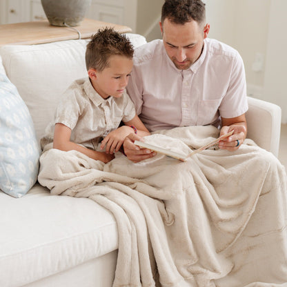 A father and son sit with a light tan lush Saranoni blanket and they are reading a book. This cozy blanket is a luxury blanket and a faux fur blanket. This soft blanket is great as a home decor staple, comfort gift, or everyday comfort item. Elevate your home decor with this faux fur blanket that is also a throw blanket. Enjoy this cozy blanket for years to come.