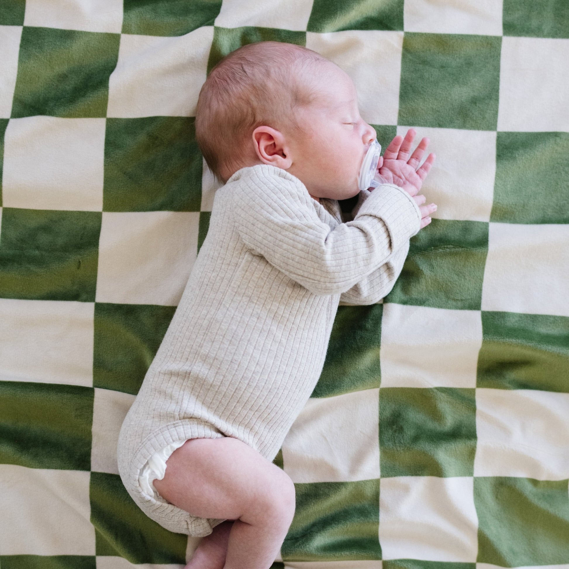 A baby boy sleeping on a olive and cream checkered minky stretch blanket. The soft blanket is a luxury blanket and a Saranoni blanket. This cozy blanket is also a receiving blanket and is a cute and cozy baby blanket. This stretchy blanket is a great nursery item. 
